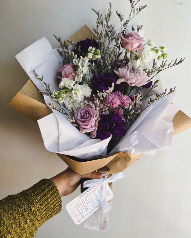 A wrapped fresh floral bouquet featuring purple flowers. Tied to it is a bag with an amethyst crystal and a card describing the metaphysical properties of amethyst.
